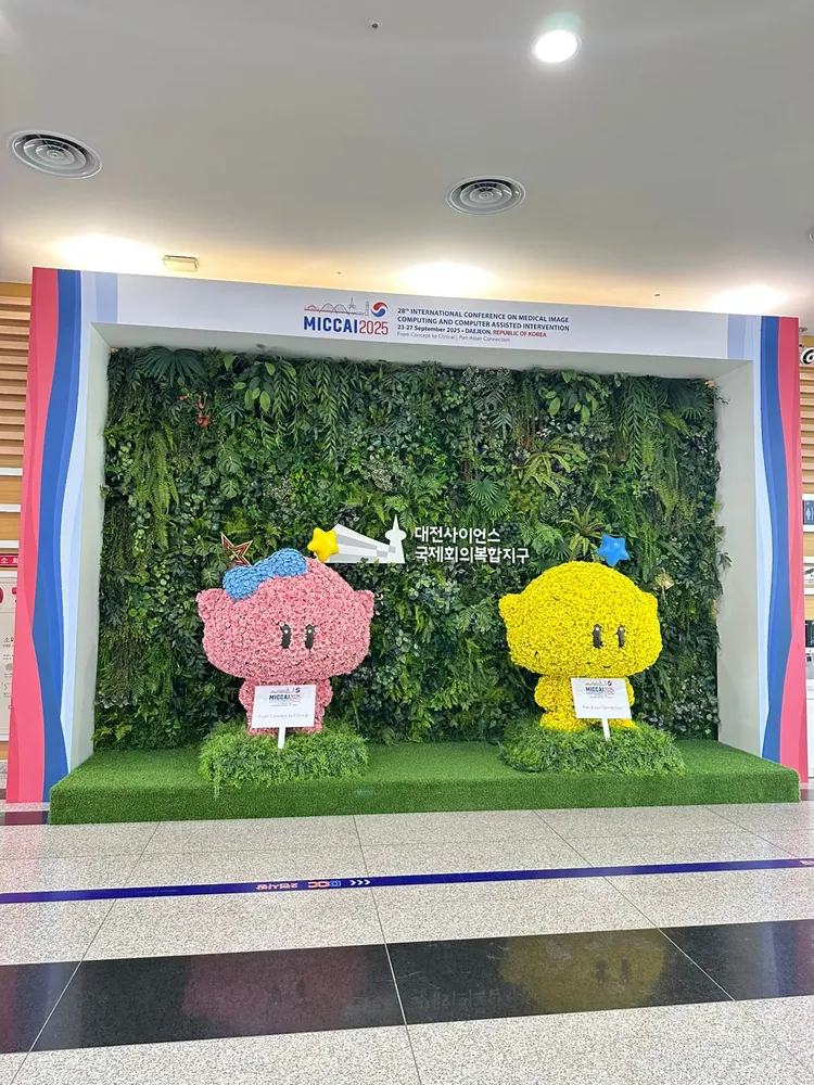 The MICCAI 2025 photo wall at the Daejeon conference venue: two flower-covered mascots, one pink and one yellow, standing on artificial grass in front of a living green wall beneath the conference banner.